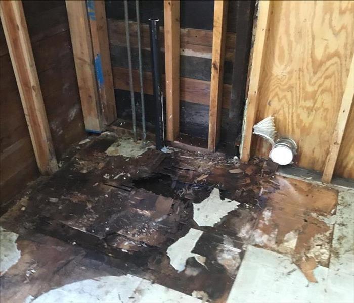 Heavily damaged subfloor near a corner with some broken flooring, heavy moisture saturation, and visible wall framing near la